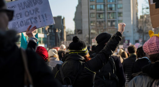 Picture of a group of people protesting in the street at a Women's March in London on 21 January 2017. They are all facing away from the camera. One woman has her fist raised defiantly in the air, and other people are holding signs and placards. Someone holds a white placard with "hands off our rights!" in big black letters.