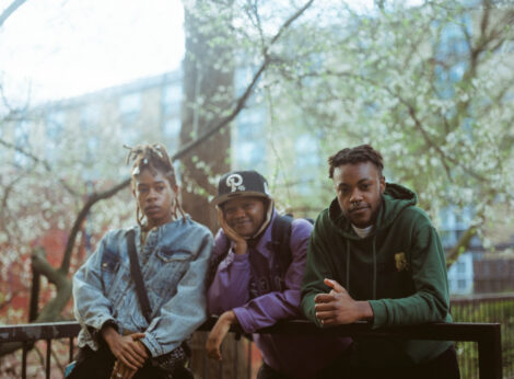 Three young black people standing next to each, one of them smiling at the camera, leaning on some railings with trees and a block of flats in the background